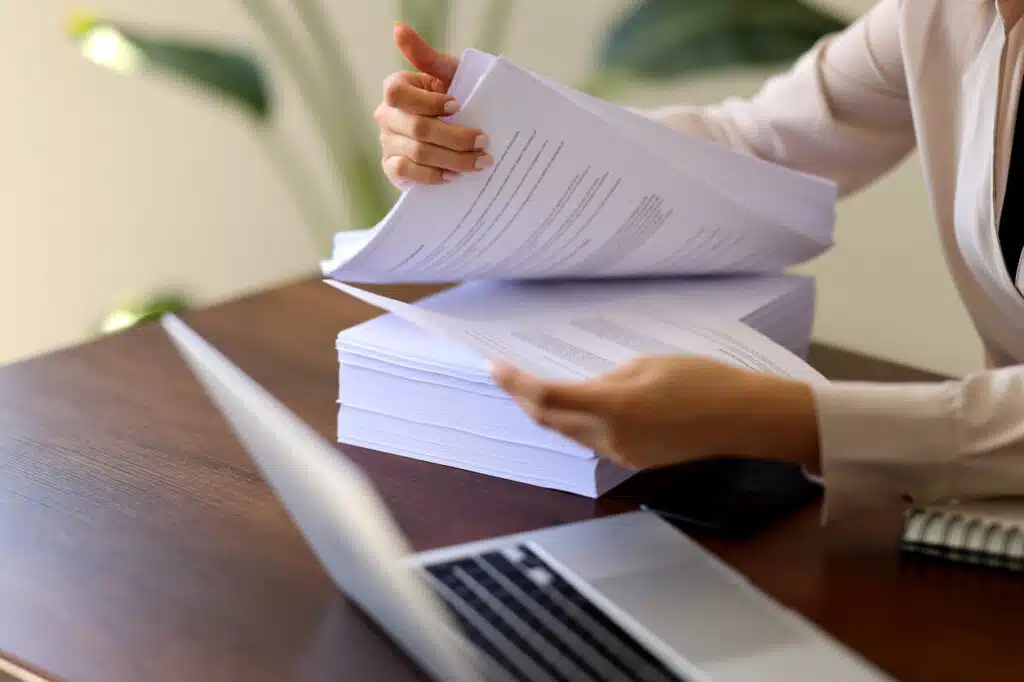 A lawyer in suit browses through a pile of legal documents while using a DMS on her computer. 